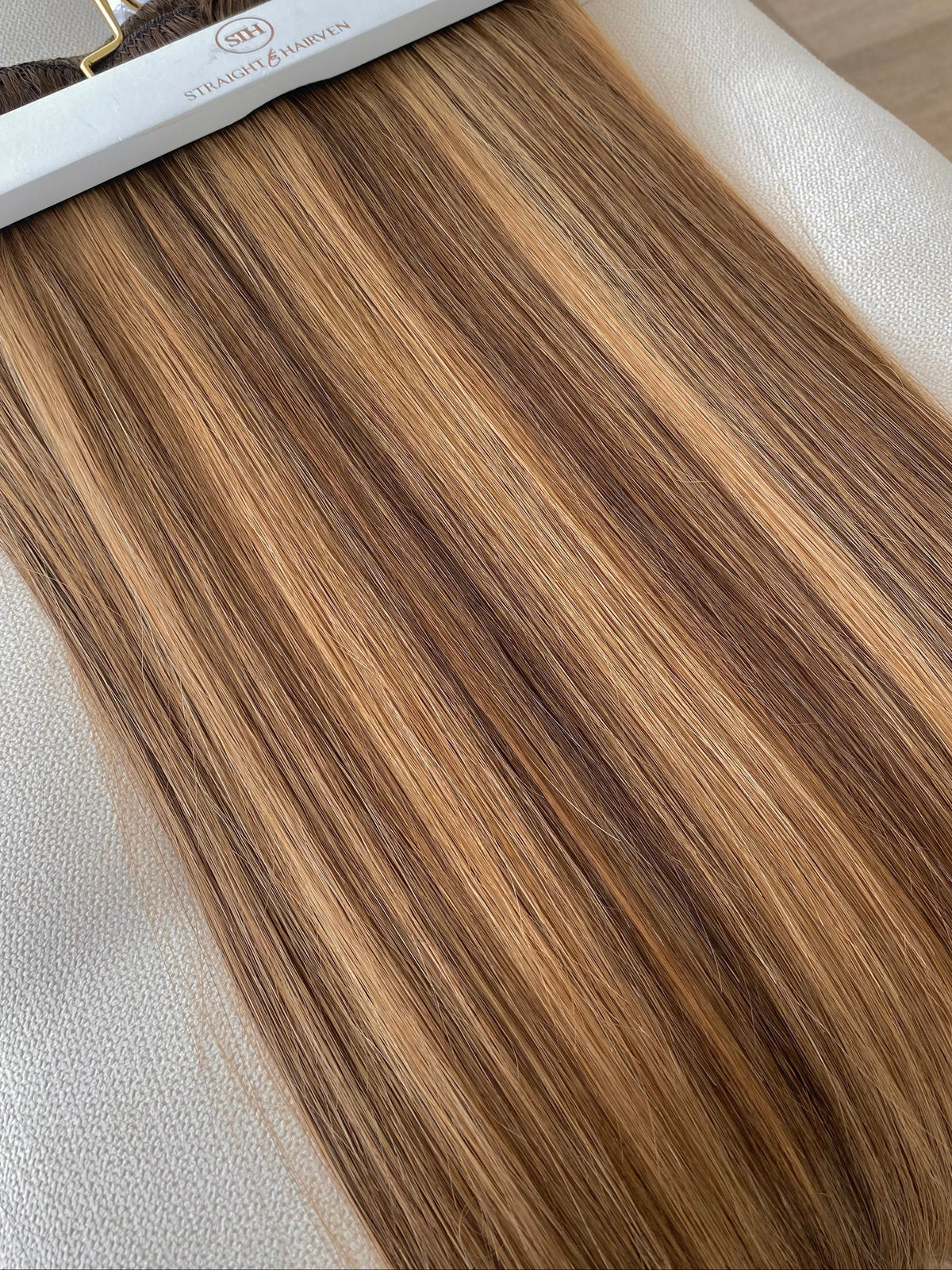 A close-up of a hair extension bundle on a white hanger, showing straight, light brown to blonde highlighted strands laid over a beige fabric surface.