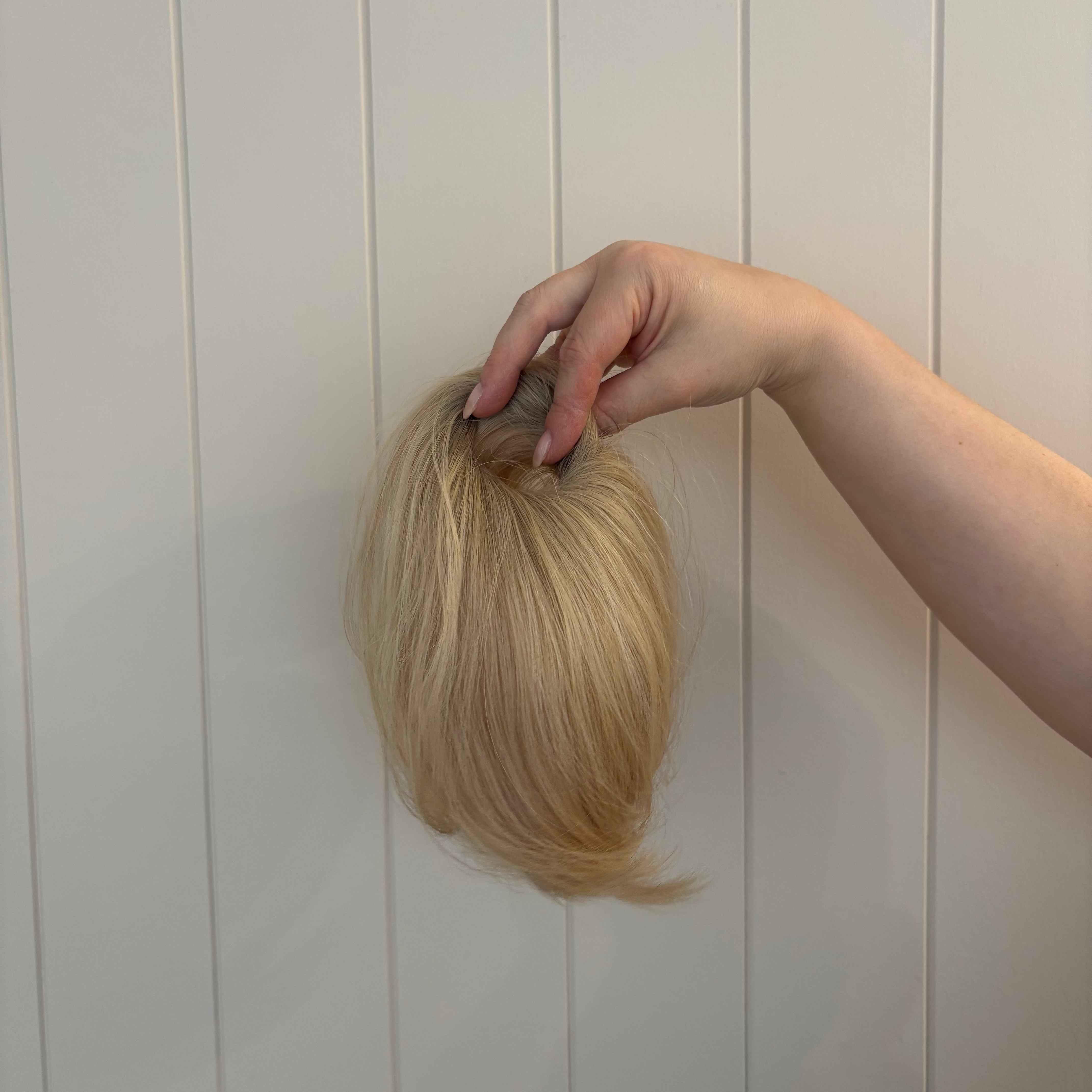 Blonde wig held by a hand against a white paneled wall