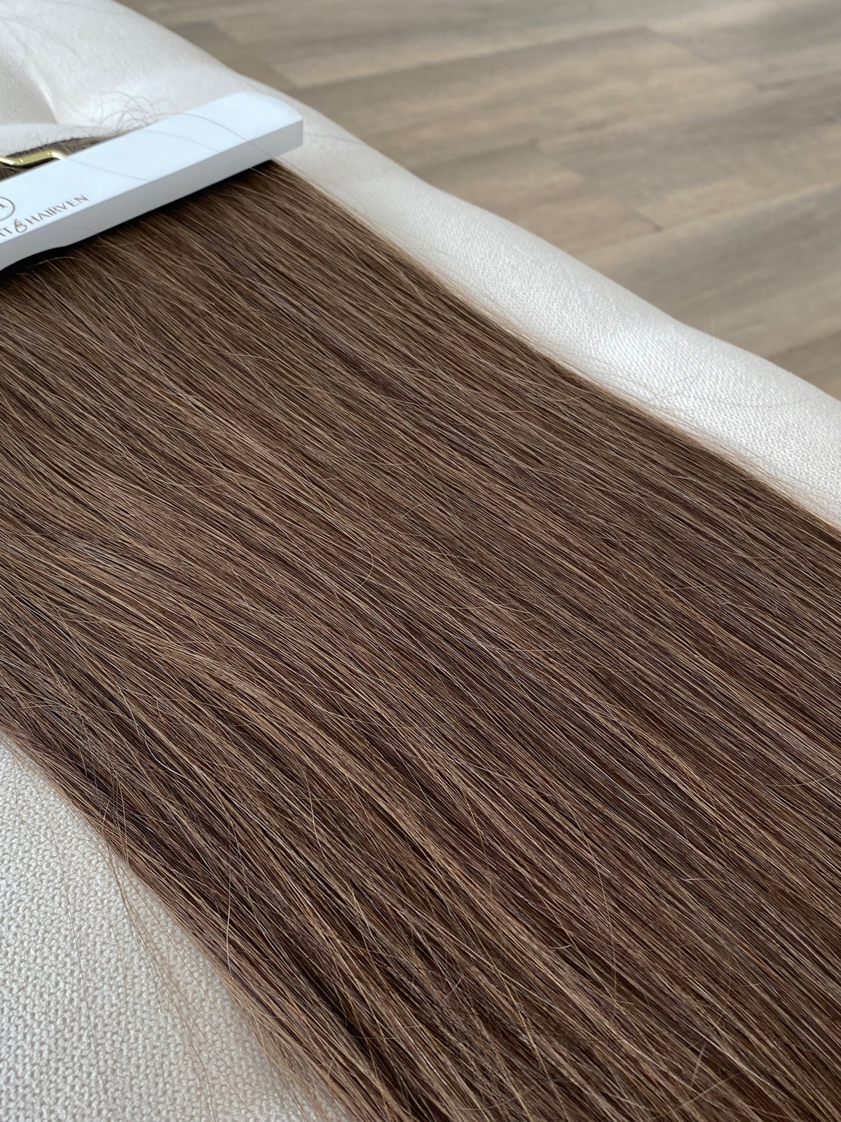 A close-up of a long, straight brown hair extension laid flat on a light beige textured surface, with part of the white retail header card visible at the top left.