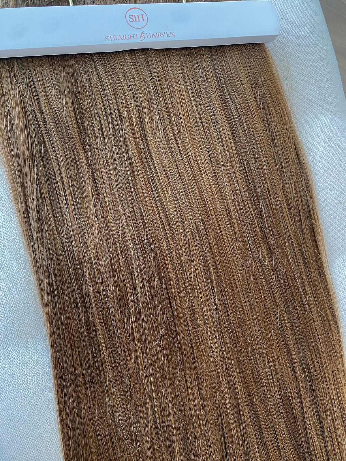 A straight hair extension swatch with light brown and golden blonde highlights, attached to a white hanger.